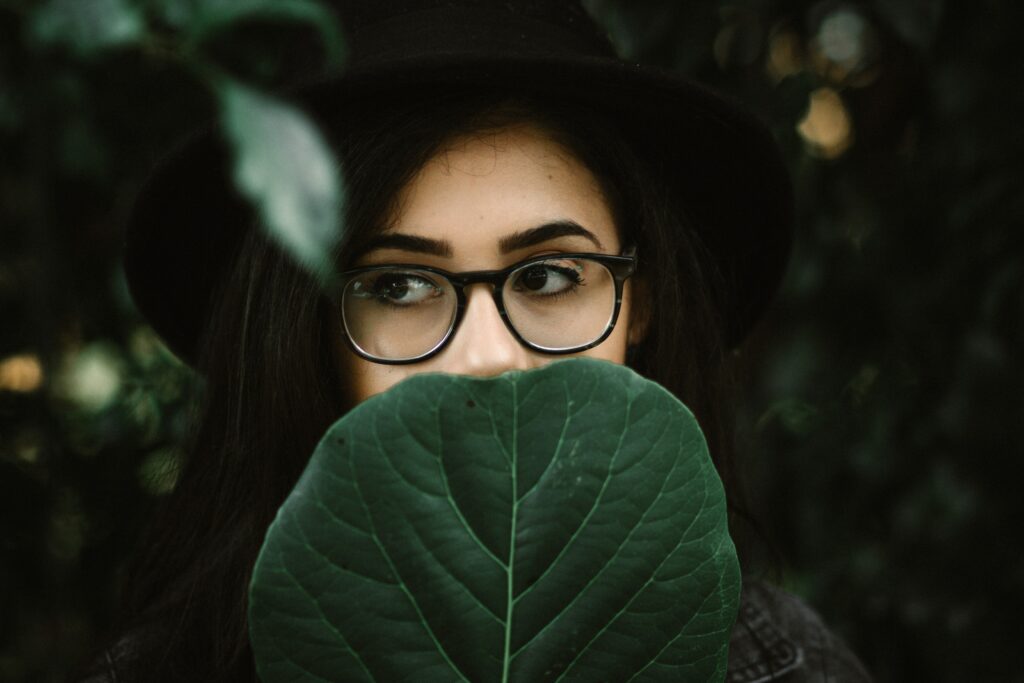 Close up of a leaf with a dark haired woman in a black hat and black rimmed glasses peeping up from behind the leaf.